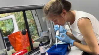 A researcher wearing blue gloves uses a pipette to transfer liquid into a small container inside a mobile laboratory van.