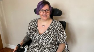 Aimee Grant is sitting on a wheelchair against a white wall. She has a short, purple hair and wearing glasses, a necklace and a black short-sleeve dress with white flower pattern. She also has tattoos on her right arm.