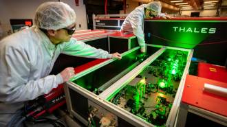 Two people in goggles and white laboratory outfits inspect optics equipment.